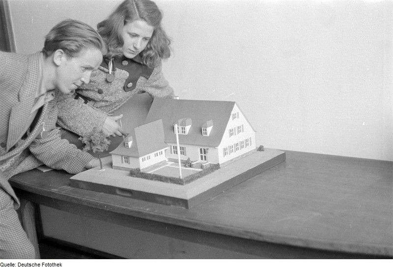 Foto: Ein altes schwarzweiss Foto. Es zeigt ein junges Paar, welches sich ein Hausmodell ansieht