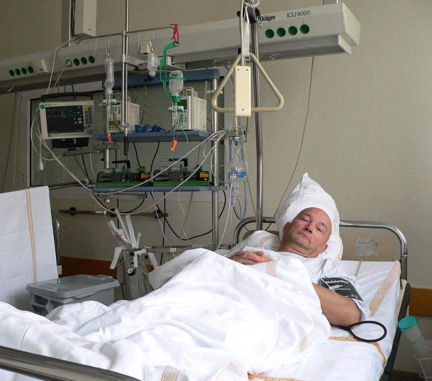 Foto: Ein kranker Patient liegt im Pflegebett im Krankenzimmer, hinter im eine Vielzahl an Kabeln und Gerätschaften