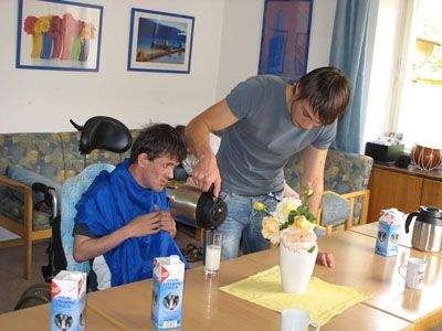 Foto: Ein Bewohner im Rollstuhl bekommt einen Kaffe eingeschenkt im sonst leeren Aufenthaltsraum