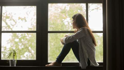 Foto: Frau hockt nachdenklich auf einer Fensterbank vor einem großen Fenster und guckt raus