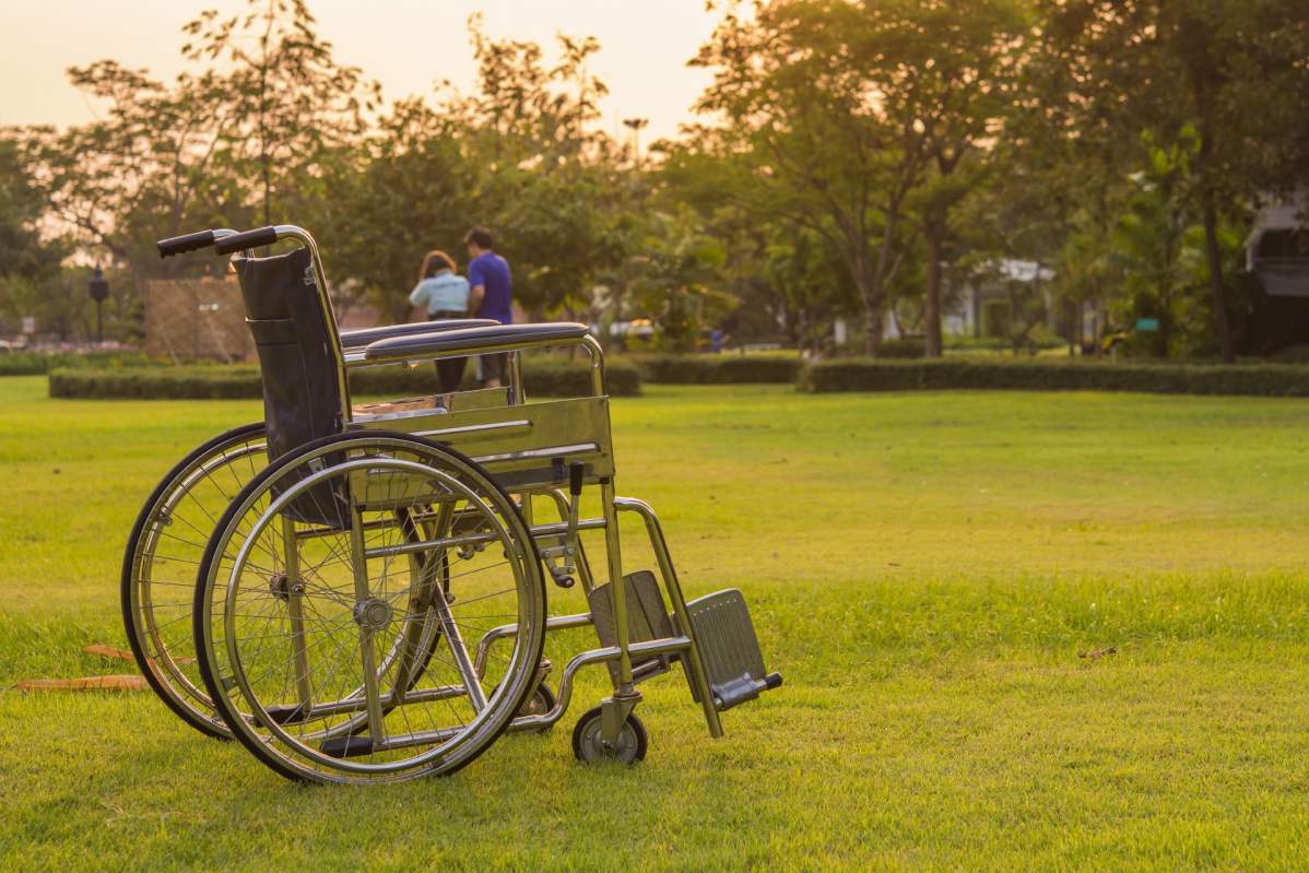 Foto: Im Vordergrund steht ein leerer Rollstuhl auf einer großen, grünen Wiese. Im Hintergrund, weit entfernt sieht man ein Paar 