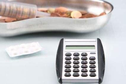 Im Vordergrund ein Taschenrechner, im Hintergrund eine Nierenschale mit Kleingeld davor ein Tablettenblister