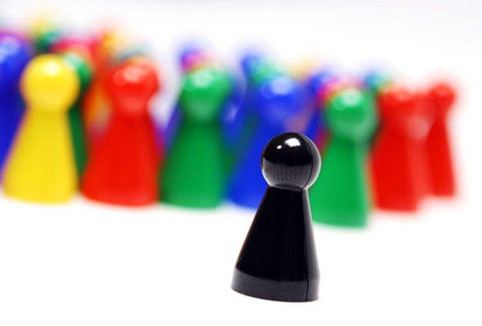 ´Viele farbige Spielfiguren stehen in einer Gruppe zusammen. Eine schwarze Spielfigur steht etwas abseits